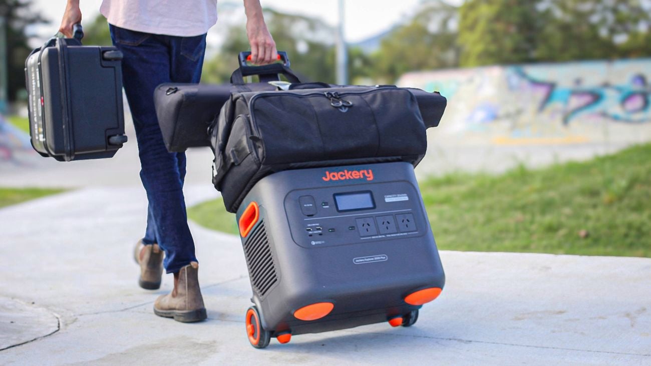 A man walks on a path carrying a case and pulling a Jackery power station with a bag placed on top.