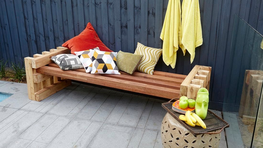 A completed outdoor day bed against a backyard fence with six throw cushions and a fruit tray nearby
