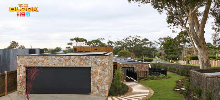 Modern garage with natural stone cladding, a black roller door, curved stepping stone pathway, landscaped lawn and mature trees.