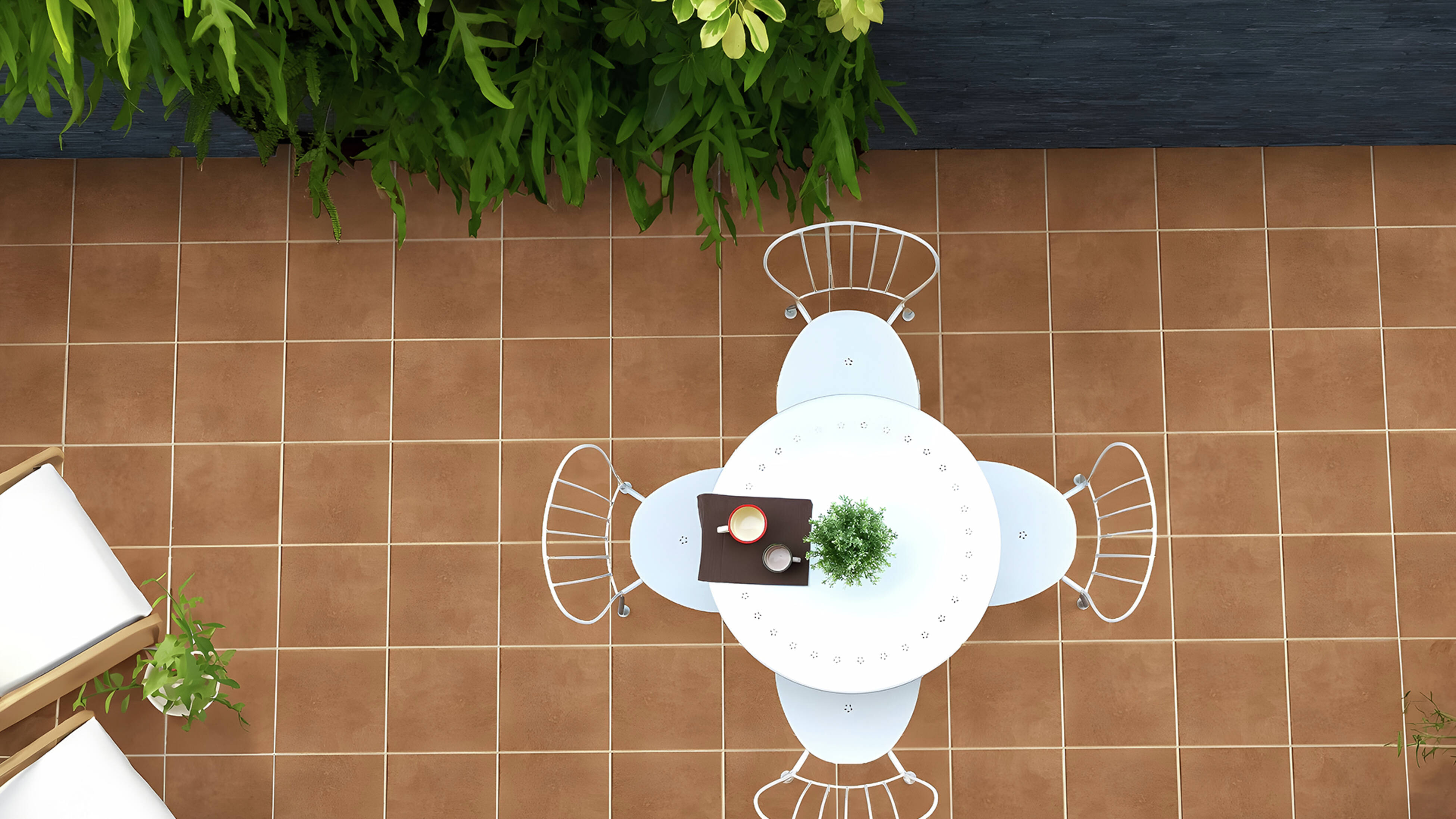 An overhead view of a patio with brown tiled flooring, a white round table with four chairs, a potted plant, and a cup. Green foliage lines the top edge, and a wooden lounge chair is partially visible.