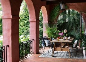 A table with crockery and a vase with flowers on it and chairs on a verandah at the back of a house.