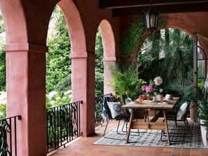 A table with crockery and a vase with flowers on it and chairs on a verandah at the back of a house.