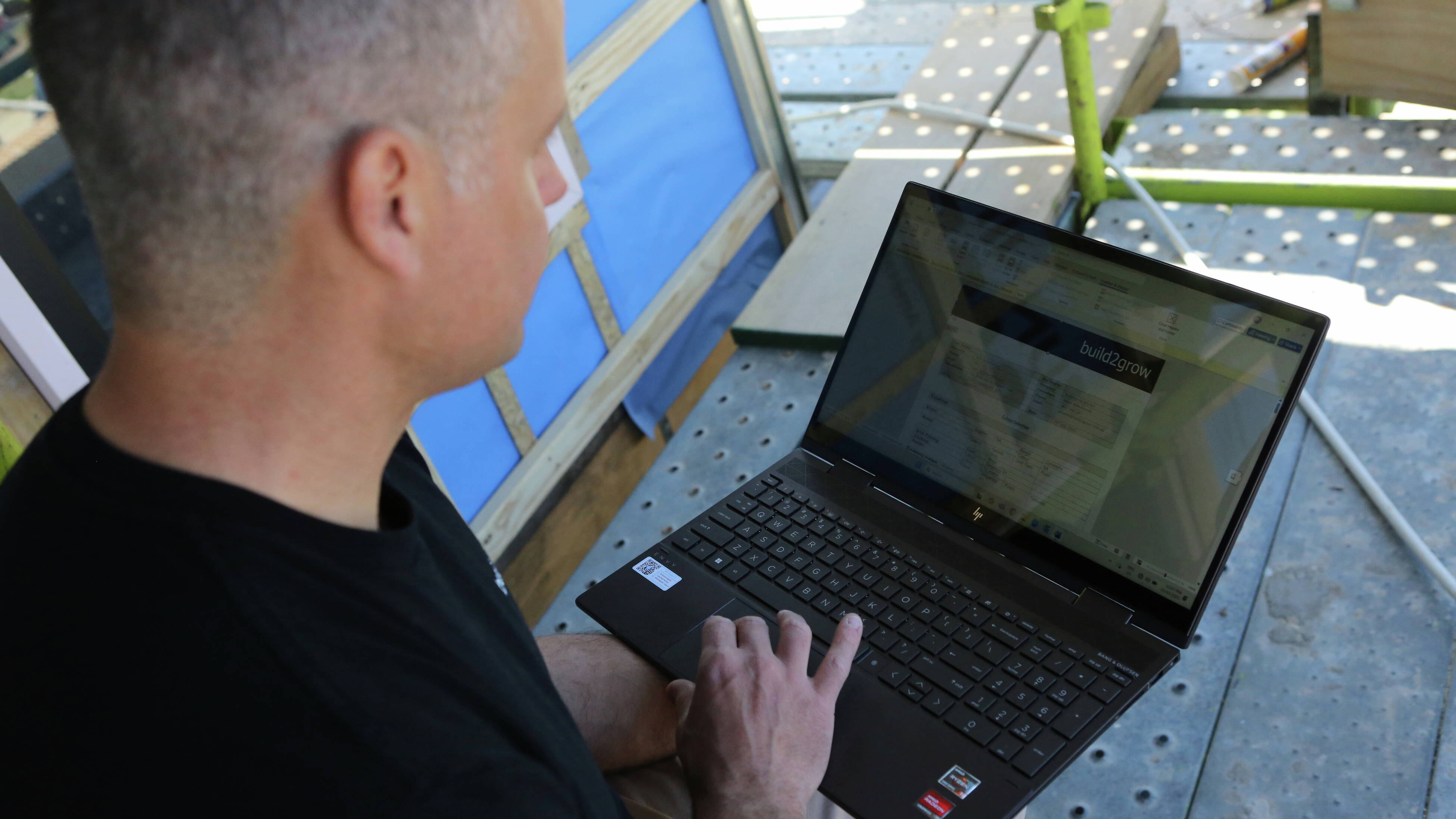 Director of build2grow Michael Nikolovski wearing a black shirt and beige pants, sits on scaffolding at a building site, holding an open laptop.