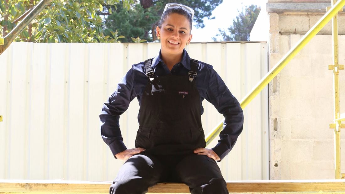A female tradesperson wearing black Green Hip overalls sits on scaffolding on a job site.