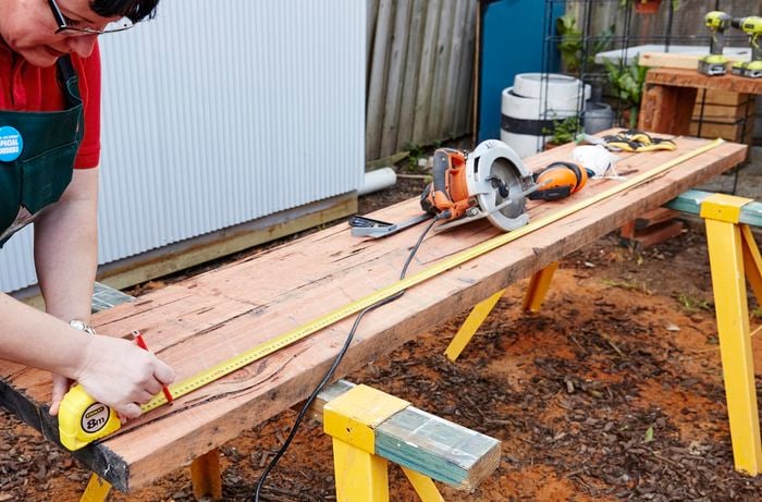 A length of sleeper being measured and marked for cutting with a tape measure and pencil, with a circular saw sitting in the background