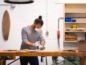 <p>Man using a rotary tool to carve wood, with 2 wooden surfboards hanging on the wall in the background.</p>
