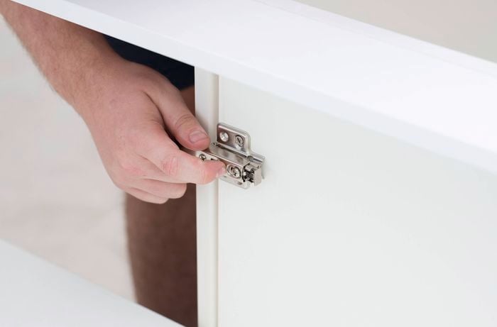 A person is attaching a cabinet door to the hinge onto a mounting plate inside a white cabinet. 