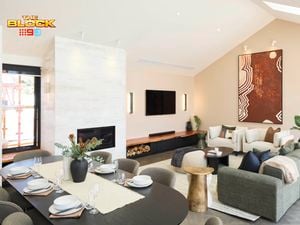 A modern open-plan living and dining area featuring a dining table set with plates, bowls, glasses, cutlery, and a floral centerpiece. The living room includes a large sectional sofa with cushions, a coffee table, and an abstract painting on the wall. A fireplace is built into a white marble wall, with firewood stored beneath a wall-mounted TV. 