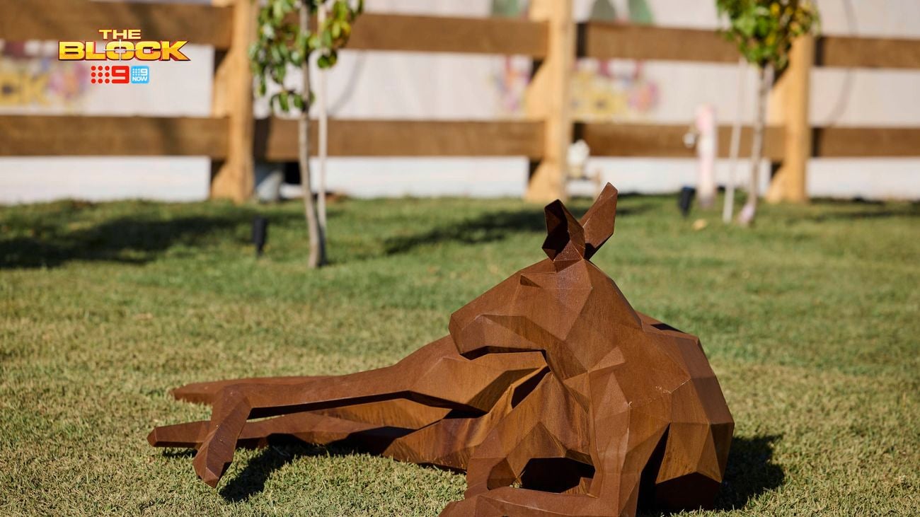 A kangaroo made out of steel laying down on a piece of grass with a timber fence around the area