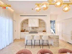 Bright, modern kitchen from The Block, featuring a marble island with four bar stools, arched range hood with stone backsplash, white cabinetry, gold fixtures, and a contemporary glass bubble chandelier.