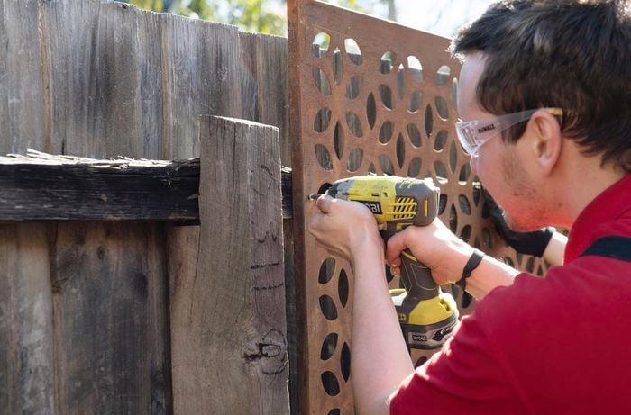 A person fixing a steel screen to a paling fence using a cordless drill
