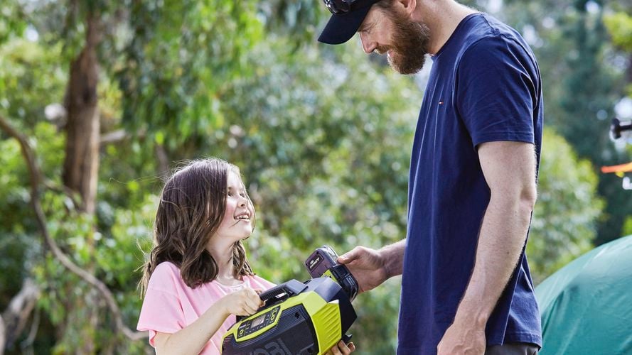 Father and daughter standing next to a tent at a campsite holding a Ryobi radio speaker with large trees in the background.