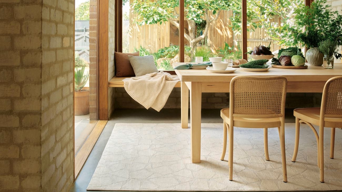 Light-filled dining area with timber table and chairs on a pale rug, set beside large windows overlooking a garden.
