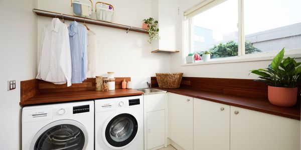 Laundry room with front-loading washer and dryer under a wooden benchtop, hanging shirts on a rail above, white cupboards, a sink in the corner, and a large window letting in natural light with indoor plants.