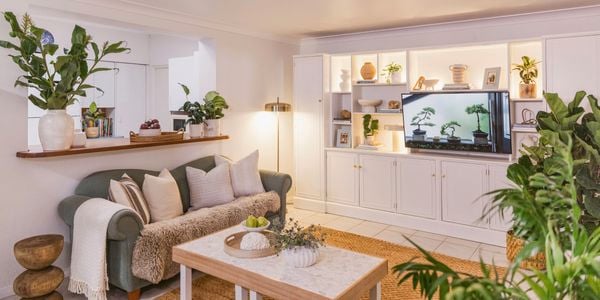 Wide view of a styled living room featuring a green sofa, coffee table, indoor plants, and white built‑in cabinetry with a television.