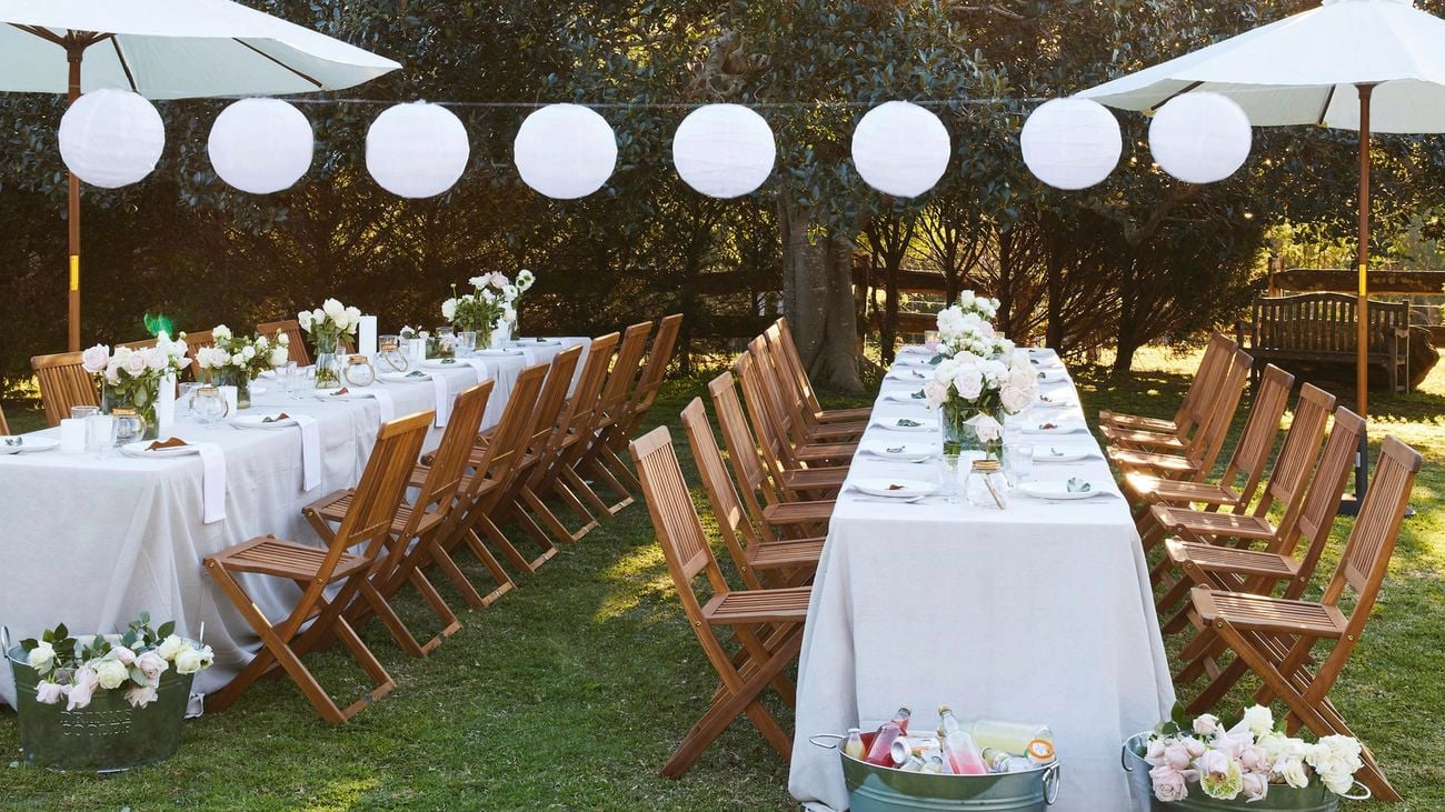 Sting of white round party lights, and two tables set for arriving guests with a white tablecloth, umbrellas and flowers.