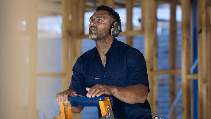A person wearing hearing protection and a navy work shirt stands on a ladder inside a timber-framed construction site.