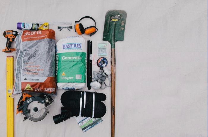 Level, drill, safety equipment, saw, shovel, and other tools and materials, laid out on the ground.
