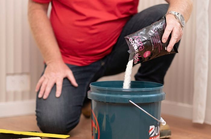 A person in a red T shirt is mixing wallpaper adhesive glue in a clean bucket.