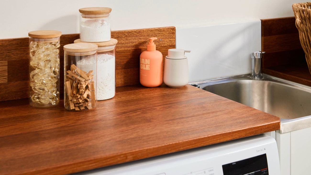 Wooden laundry benchtop with glass storage jars, cleaning liquids in pump bottles, and a stainless steel sink set into the corner, against a white wall with timber backsplash.