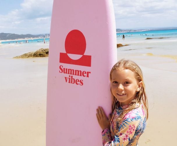 Smiling young girl standing behind a pink Summer vibes soft surfboard on a sunny beach.