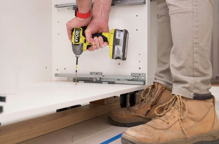 A person is using a power drill to secure the base of a cabinet to the timber support frame.