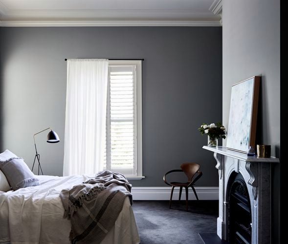 Dark tone bedroom with ornate fireplace and blanket draped on bed in front of a window with plantain shutters and a sheer curtain