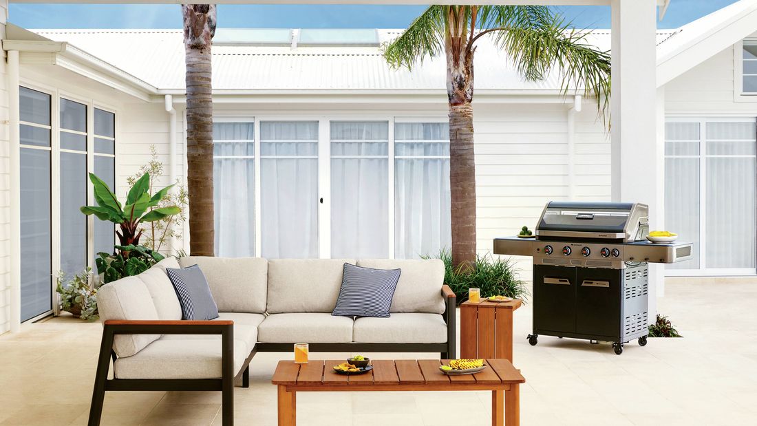 Covered outdoor entertaining area featuring a cushioned sofa, timber coffee table, barbecue and palm trees.