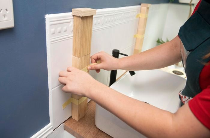 A person taping blocks of wood to a tiled wall above a basin in a bathroom