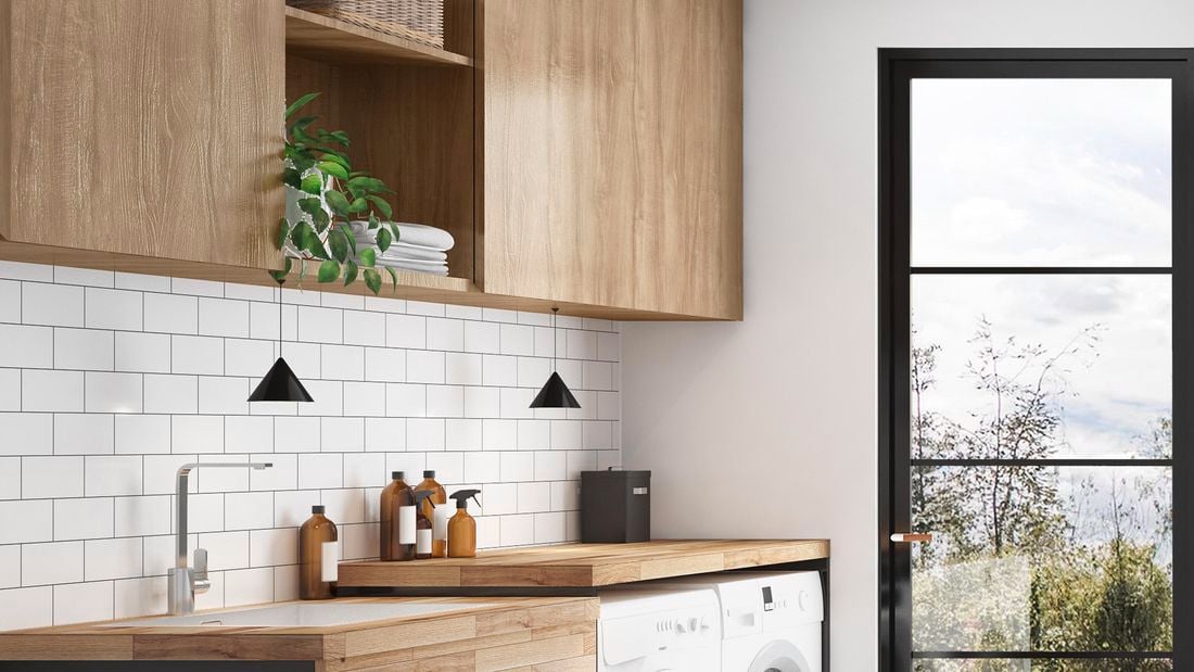 A pale white and wooden laundry area with overhead cabinets, wooden benches and whitegoods, and a black windowed door overlooking trees