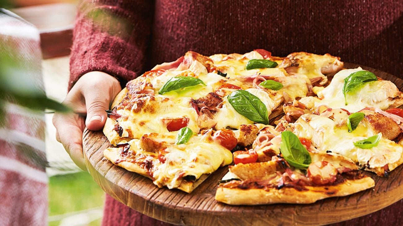 A person holding a chicken pizza on a timber serving board