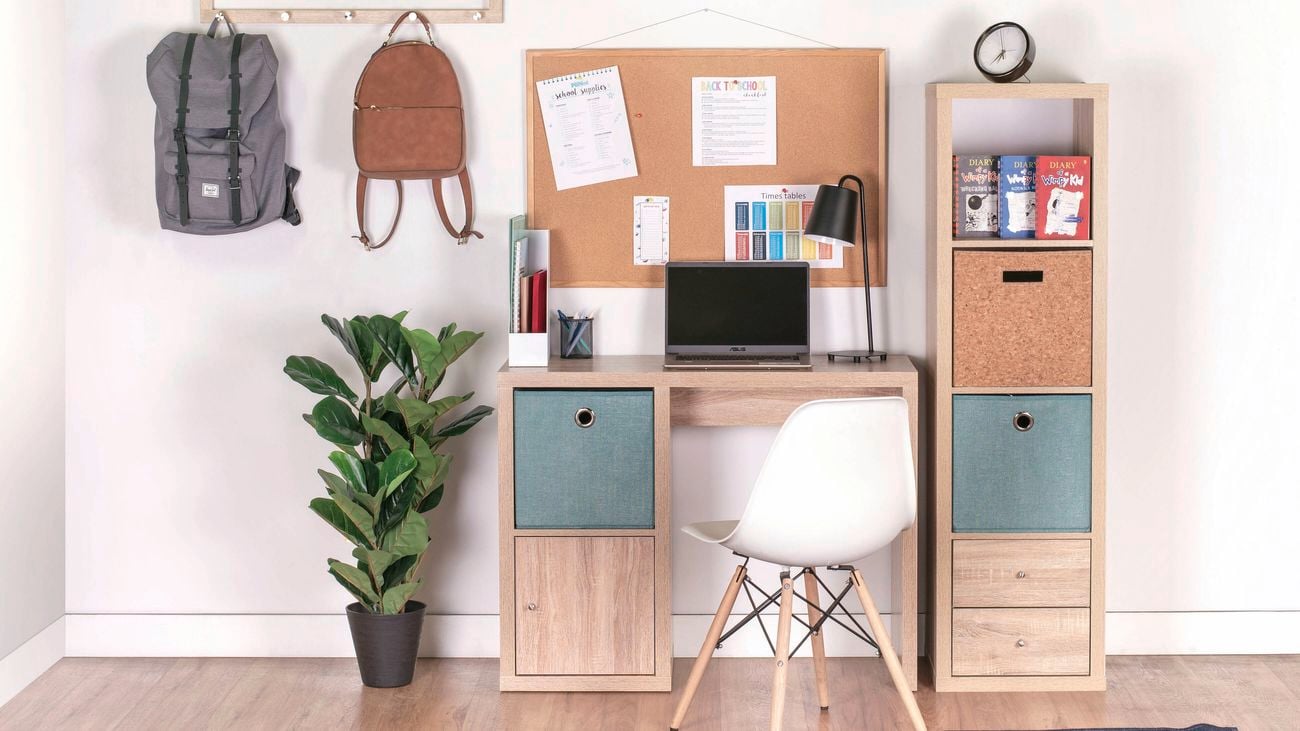 A desk, corkboard, chair, shelving unit, plant on a timber floor.