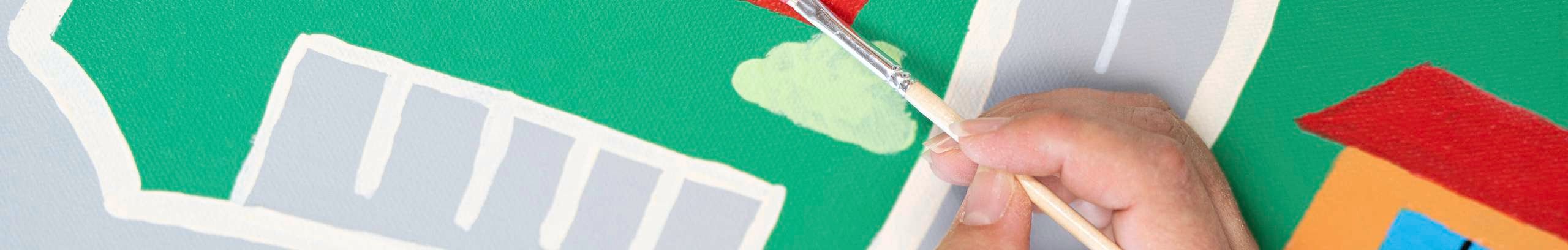 A hand painting a red house on a colourful mural map showing roads, a shop, and a school.