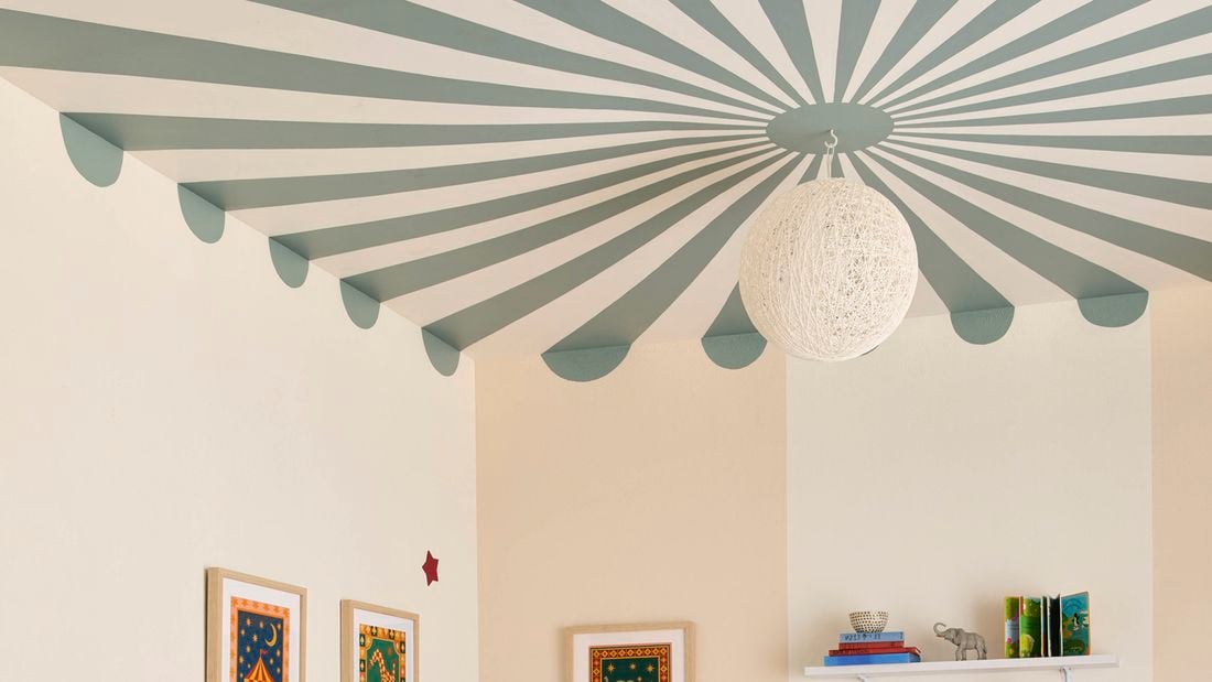 Ceiling mural painting with green and white radial stripes and a scalloped edge in a room corner, with a pendant light, cream walls, framed artwork and a bookshelf.
