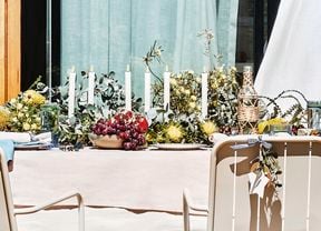 Outdoor table setting with a light tablecloth, a long greenery centrepiece holding white taper candles, and bowls of grapes and other fruit. Sliding glass door with pale curtains behind; beige chairs, one tied with a ribbon and foliage, and a twine‑wrapped bottle on the table