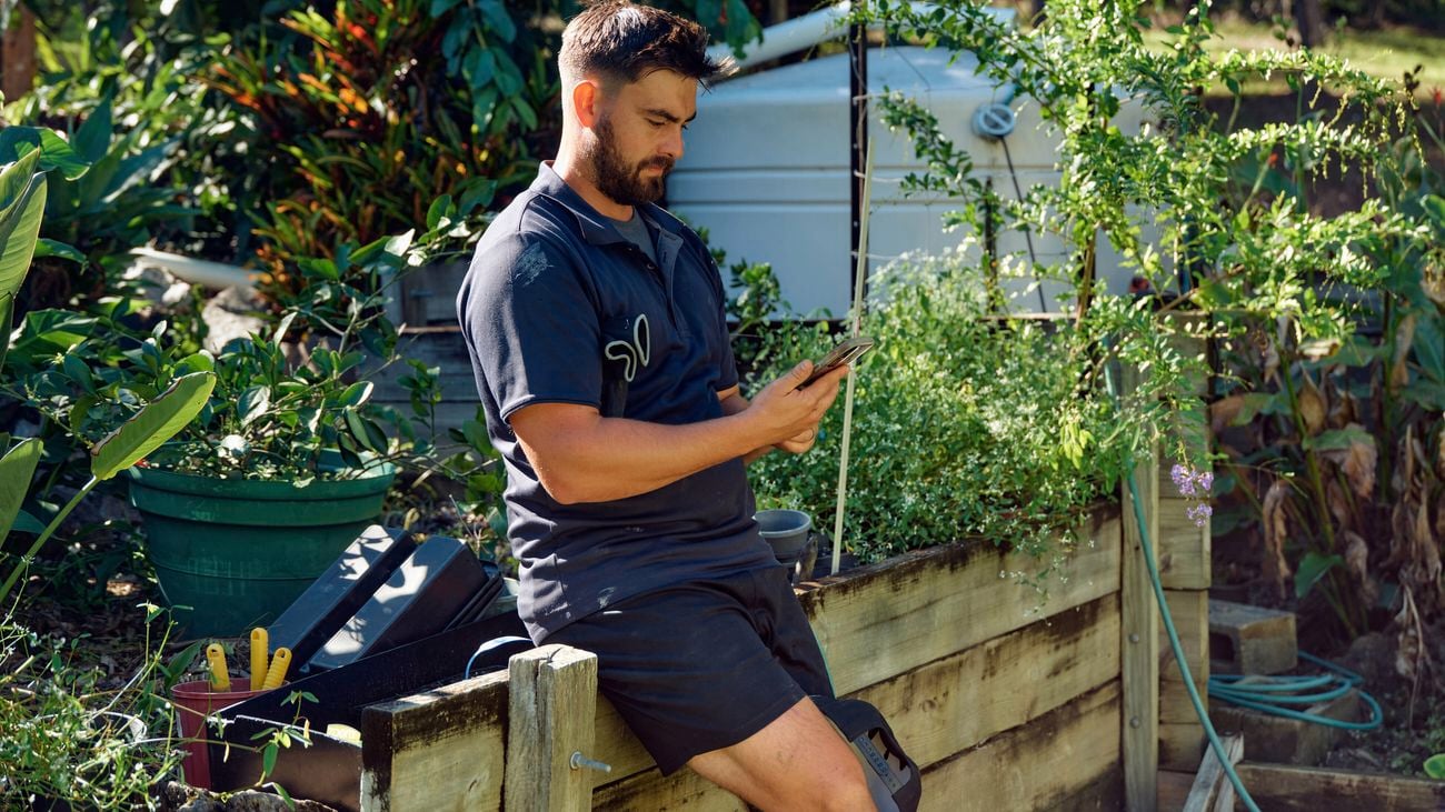 A man in a blue shirt sits on a retaining wall in a garden looking at the Bunnings Trade PowerPass App on his phone.