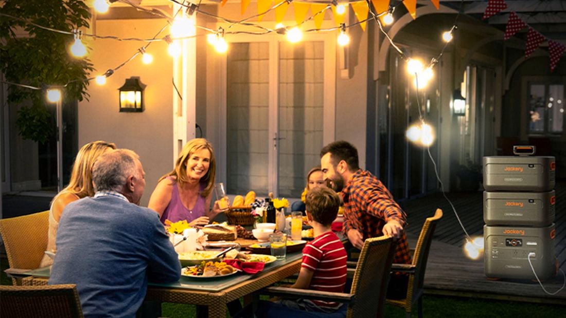  A group of people seated around an outdoor dining table on a verandah at night eating dinner with festoon lights above and a Jackery Power Station in the background.