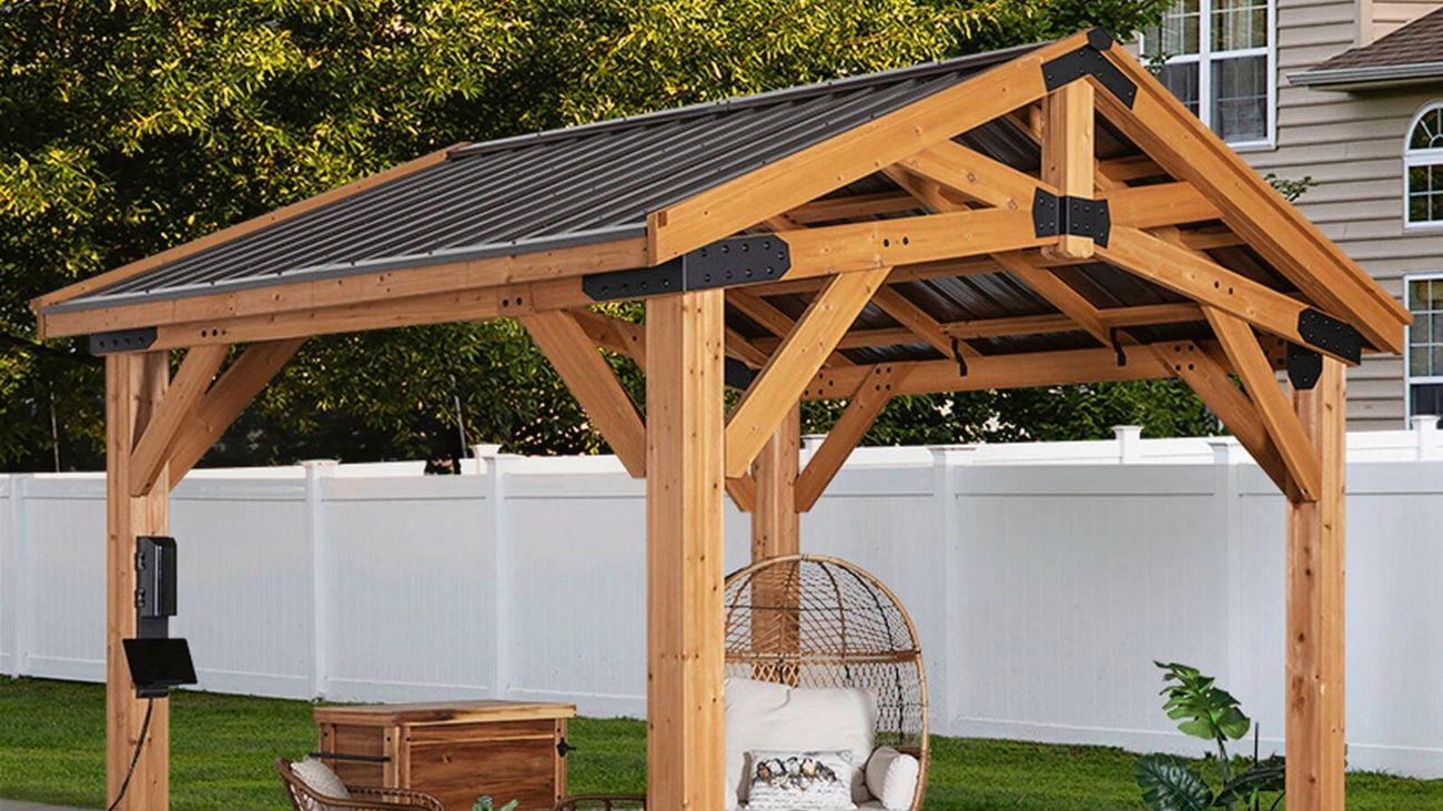 A wooden gazebo with a dark roof covering an outdoor patio with a hanging egg chair, a small table with two chairs, and a wooden storage box.