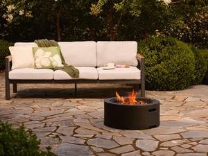 Low black outdoor fire pit with a visible flame, placed on a stone paved patio in front of a modern outdoor sofa and landscaped garden.