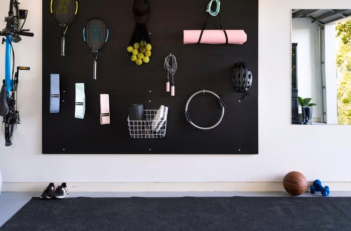 A gym area in a garage featuring a wall mirror, wall mounted bike and plywood panels used as wall storage for gym and yoga equipment