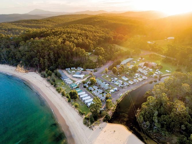 Aerial view of a coastal campground with rows of caravans near a sandy beach and clear blue water, surrounded by dense forest and hills under a warm sunrise or sunset glow.