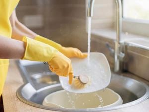 A woman wearing yellow dishwashing gloves and a yellow apron uses a scrubbing brush to wash dishes in a sink