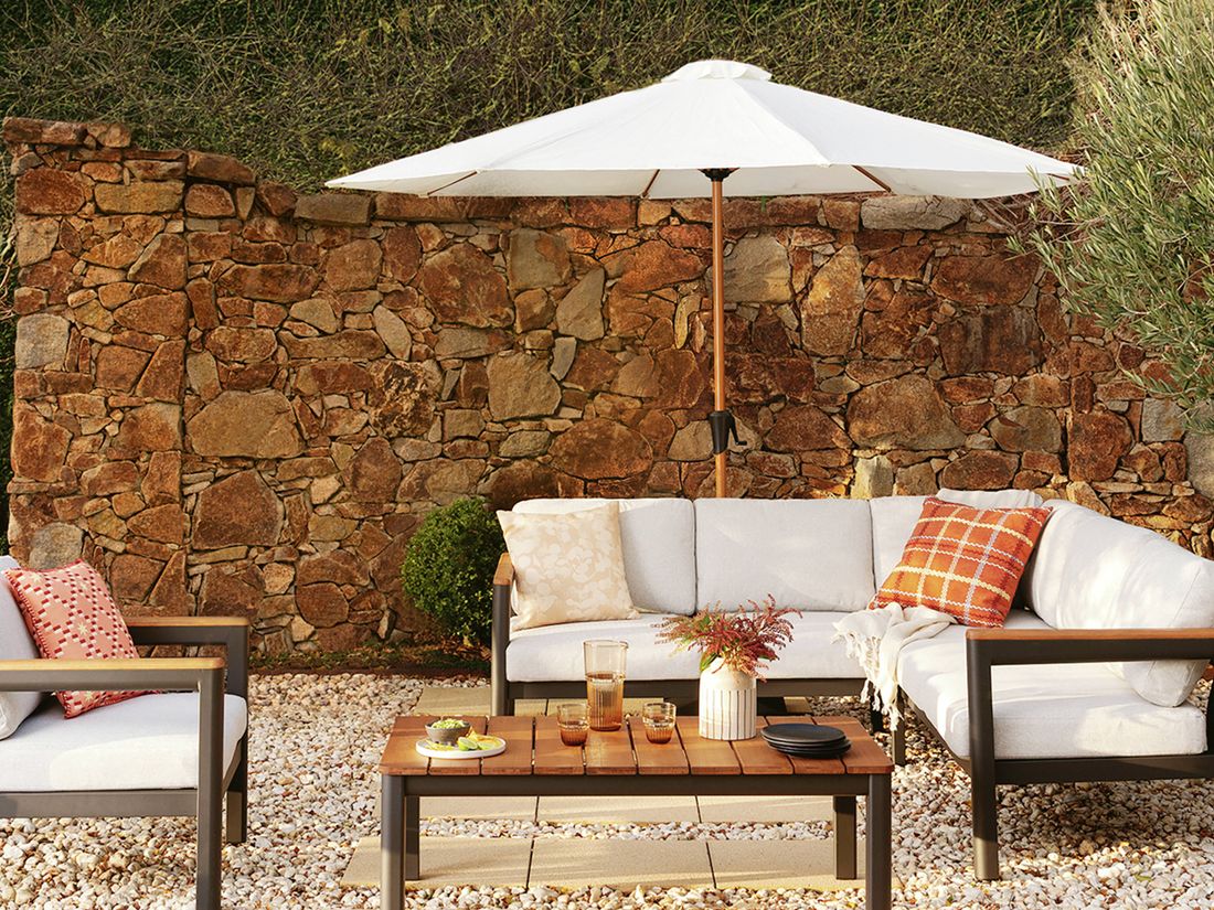 : Outdoor patio with an L-shaped lounge, armchair, and coffee table under a white umbrella, against a stone wall and hedges