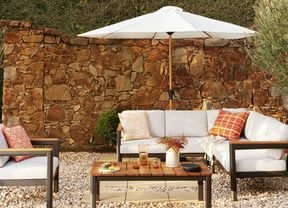 : Outdoor patio with an L-shaped lounge, armchair, and coffee table under a white umbrella, against a stone wall and hedges