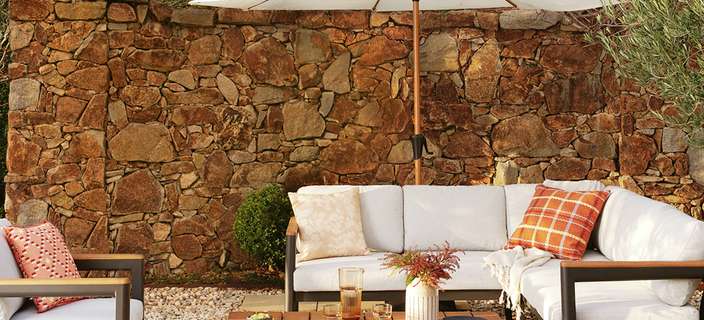: Outdoor patio with an L-shaped lounge, armchair, and coffee table under a white umbrella, against a stone wall and hedges