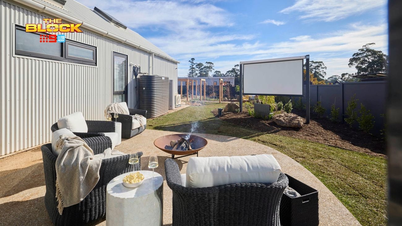 An outside area next to a house, with an outdoor setting, a fire pit, portable screen and a grassed area in it.