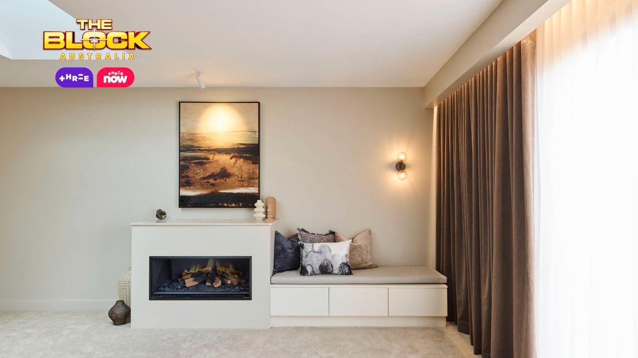 A spacious, modern bedroom with a large bed dressed in a plaid blanket and pillows. The room includes a skylight, a wooden accent wall, a built-in fireplace with a seating area, and large windows with curtains that allow natural light to enter.