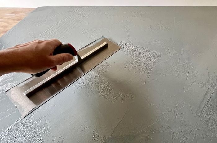 A door panel covered in wet grey paint, with a hand holding a metal trowel wiping it over the paint.