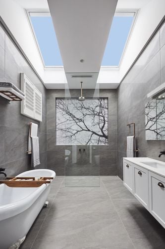 Long bathroom with grey floor and wall tiles, white vanity and freestanding bathtub and large skylight