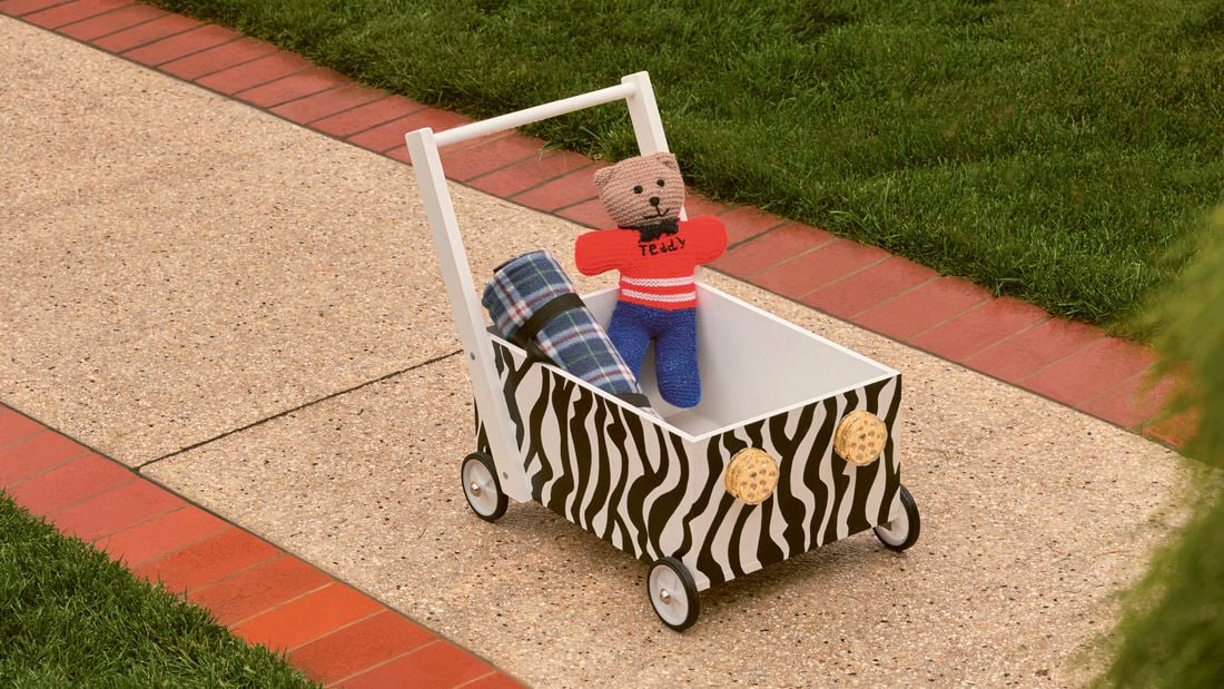 : A zebra-print toy wagon with a teddy bear and a plaid blanket on a paved path next to a lawn.